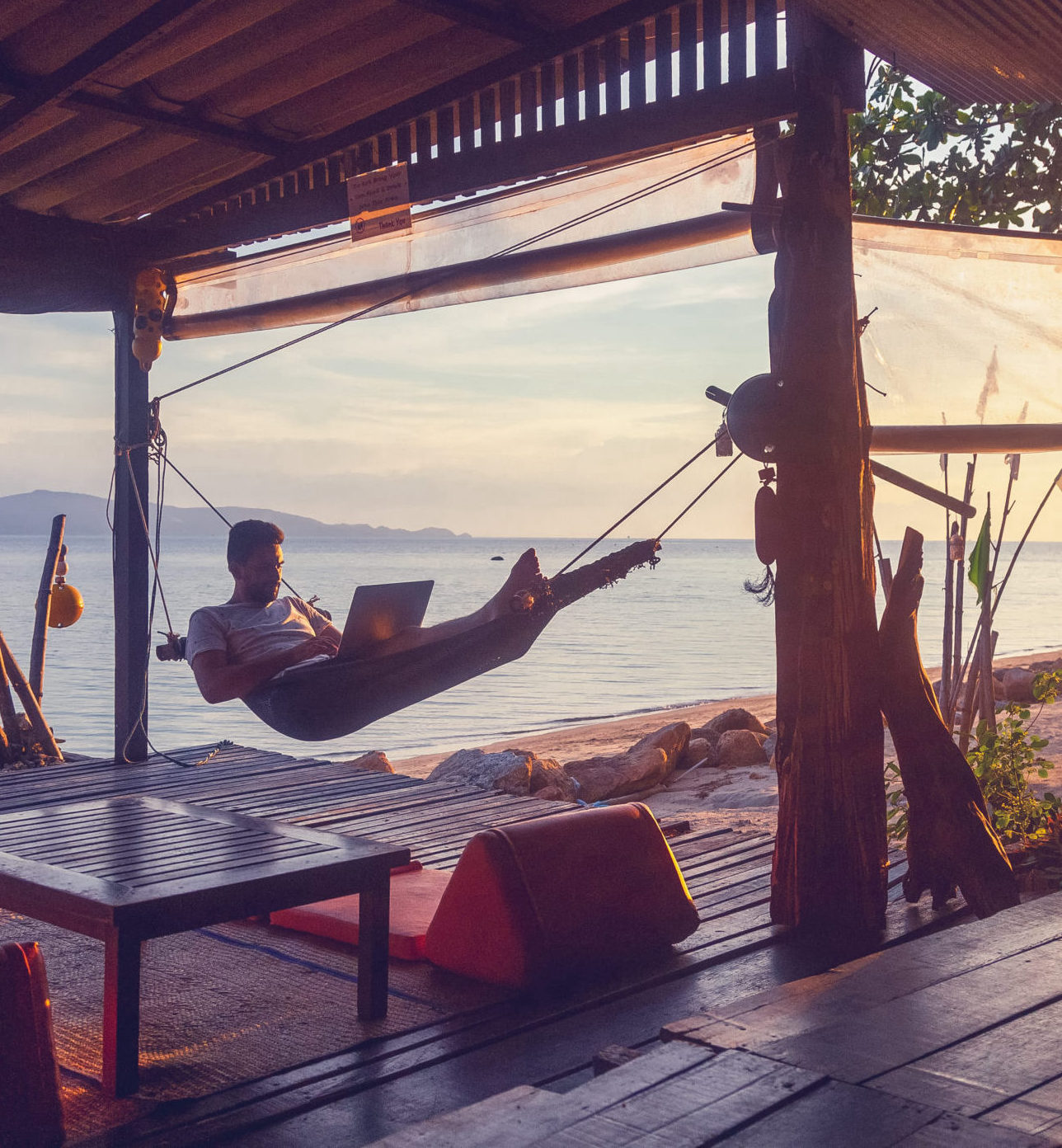 Remote Work in Paradise: Hammock with a View A man working on a laptop while relaxing in a hammock under a wooden canopy, overlooking a tranquil seaside view at sunset.