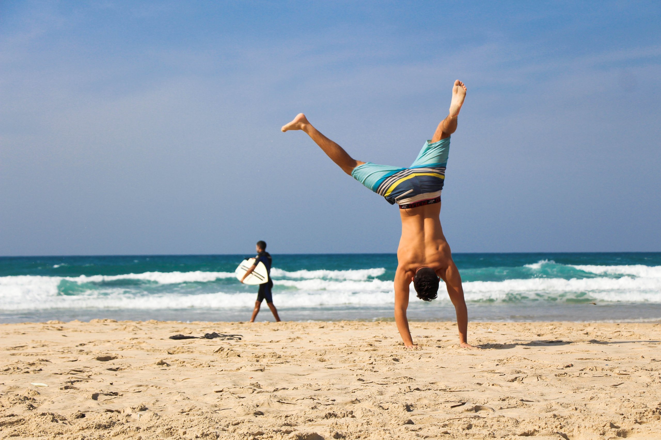 A man doing a handstand on a sandy beach with the ocean in the background, while another person walks nearby holding a surfboard.