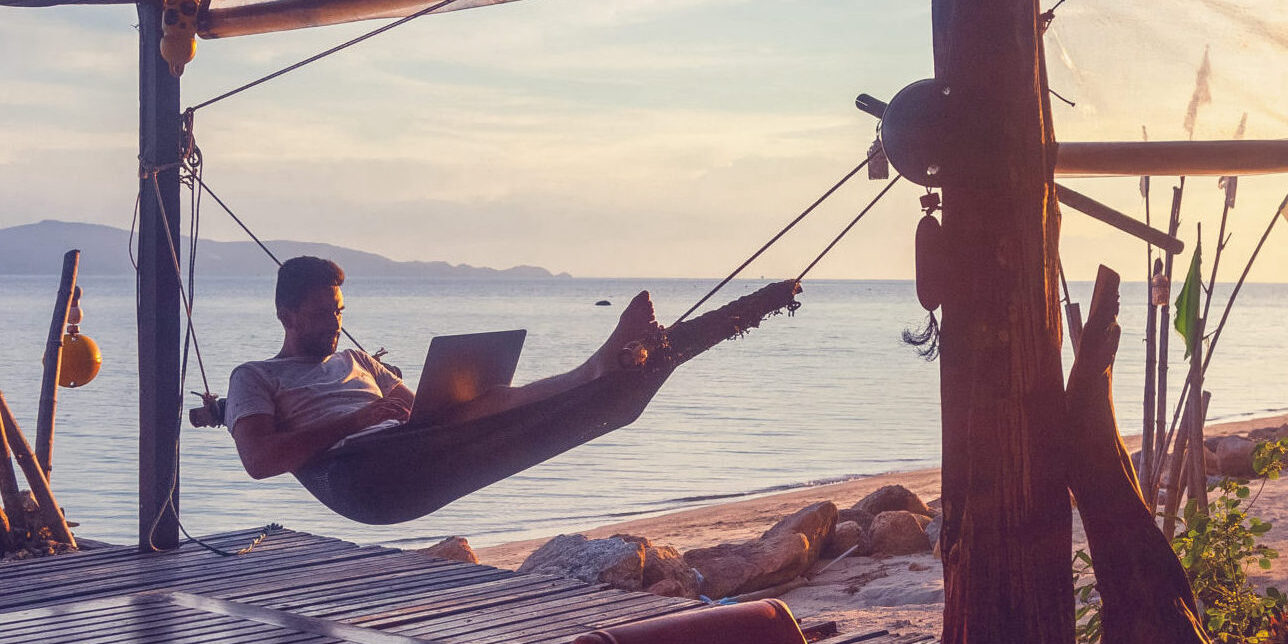 Remote Work in Paradise: Hammock with a View A man working on a laptop while relaxing in a hammock under a wooden canopy, overlooking a tranquil seaside view at sunset.