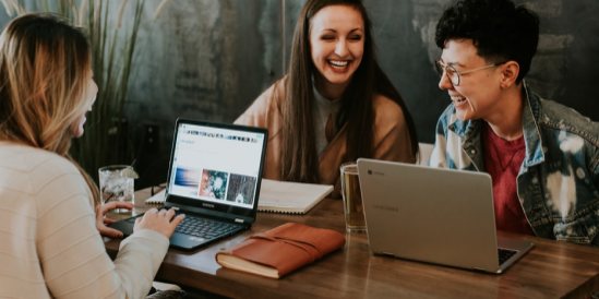 Collaborating for Success: Building Income Streams Together Three people laughing and collaborating with laptops at a wooden table, symbolizing teamwork and creating income streams online.